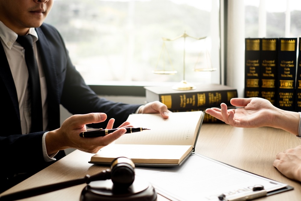 A criminal defense attorney is consulting with a client in a law office, reviewing legal documents with a pen, law books, and a judge’s gavel to discuss legal strategy and protect the client’s rights.